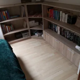 Kątowa zabudowa regałowa z jasnego drewna, wypełniona książkami, pudełkami i dekoracjami, z widocznymi frontami szafek w dolnej części i fragmentem zielonego pledu w lewym dolnym rogu.