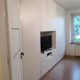 Biała zabudowa meblowa pod telewizor z szafkami i półkami, widok z boku, w tle okno z widokiem na zieleń.