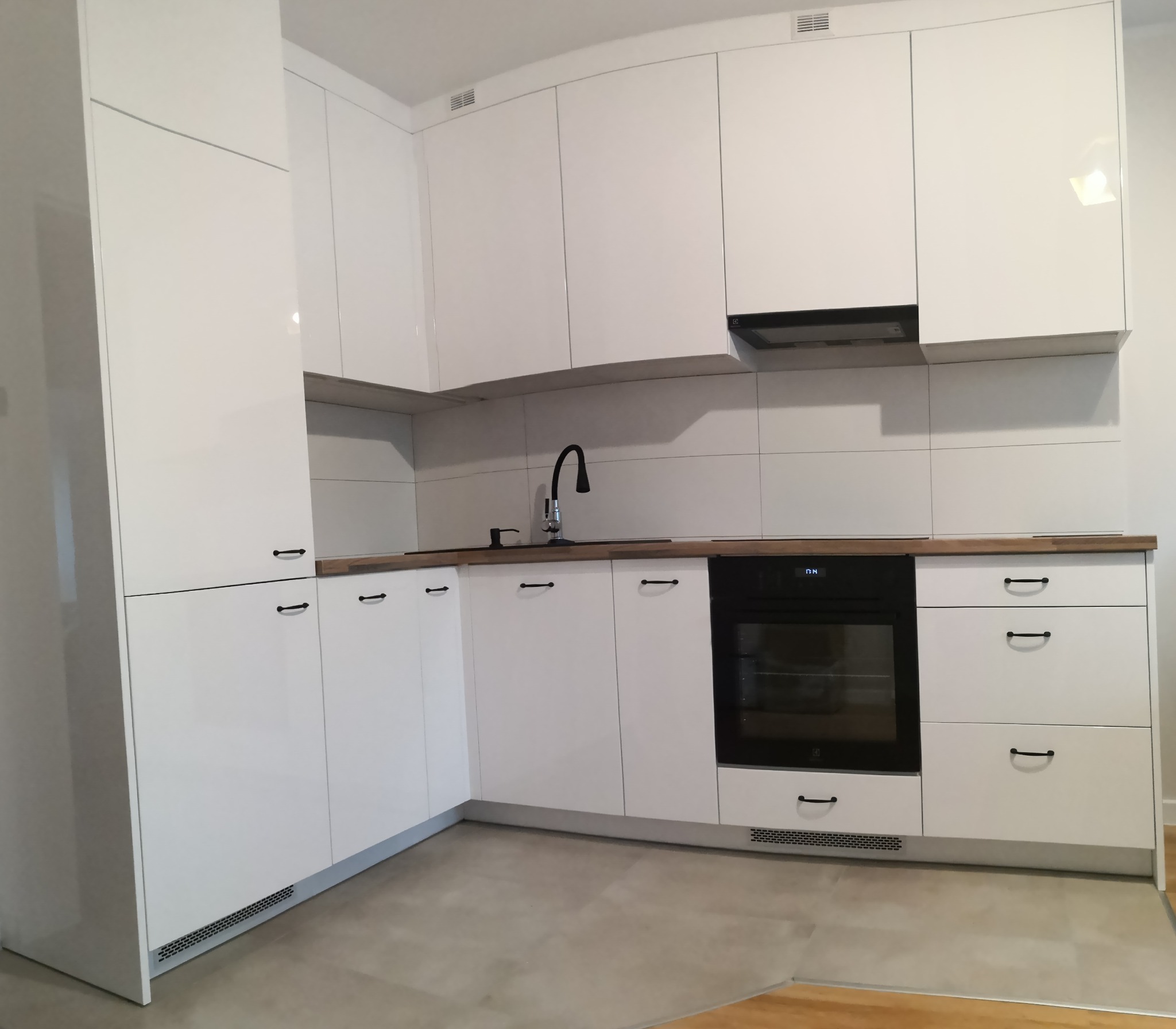 Nowoczesna, minimalistyczna kuchnia z białymi, lakierowanymi frontami, czarną armaturą i drewnianym blatem, widok na piekarnik i szafki.