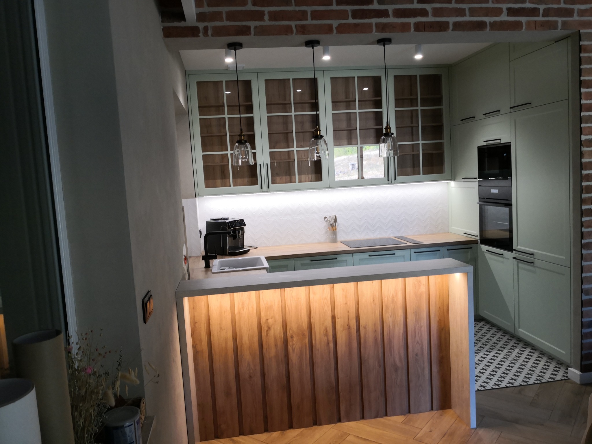 Jasna kuchnia z drewnianym blatem barowym podświetlonym od dołu, wiszące lampy nad blatem, ekspres do kawy na blacie, wzorzysta mozaika na podłodze.