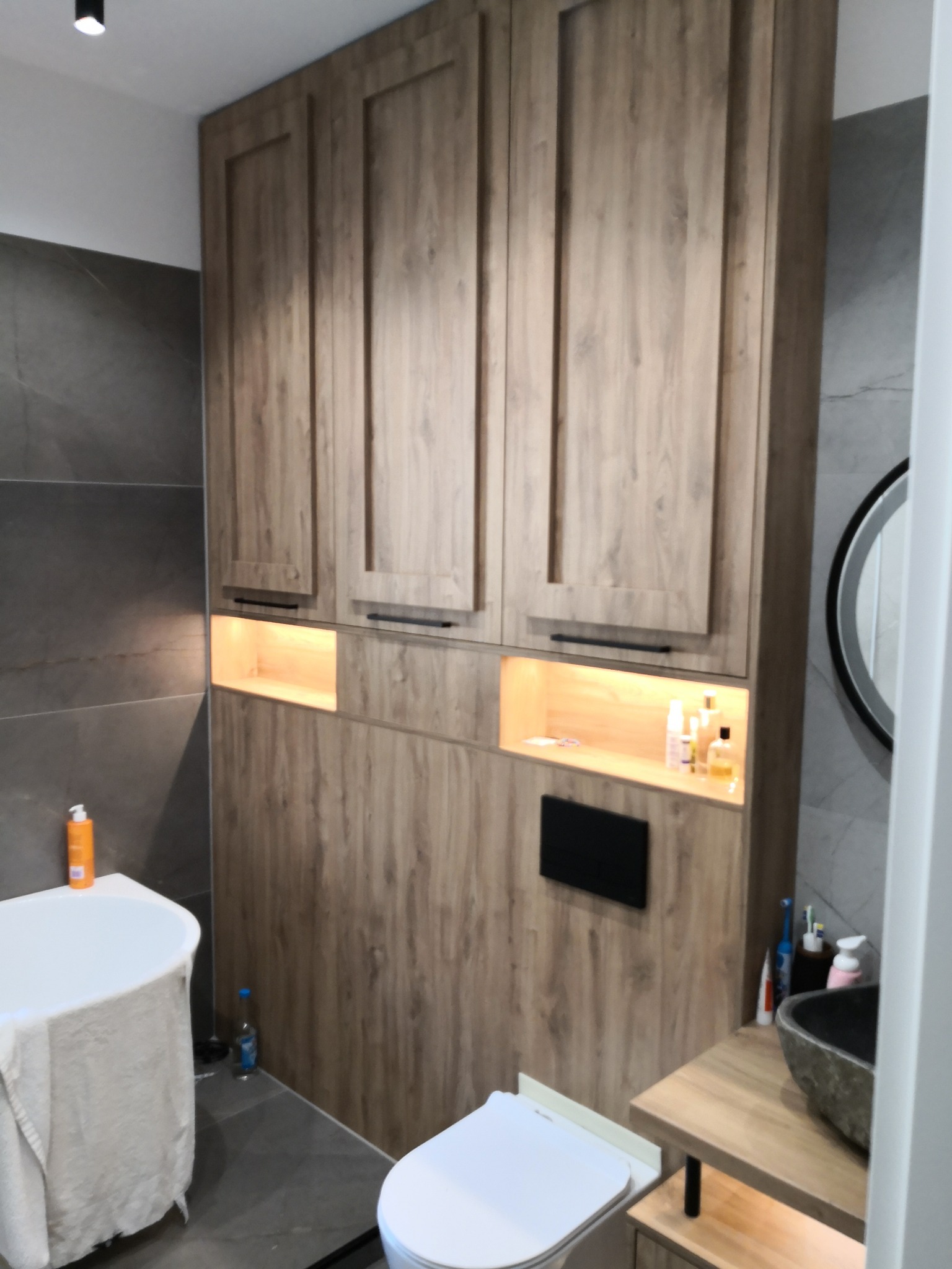 Drewniana zabudowa w łazience z szafkami, oświetlonymi wnękami, umywalką z kamienną misą i toaletą, na tle szarych płytek.