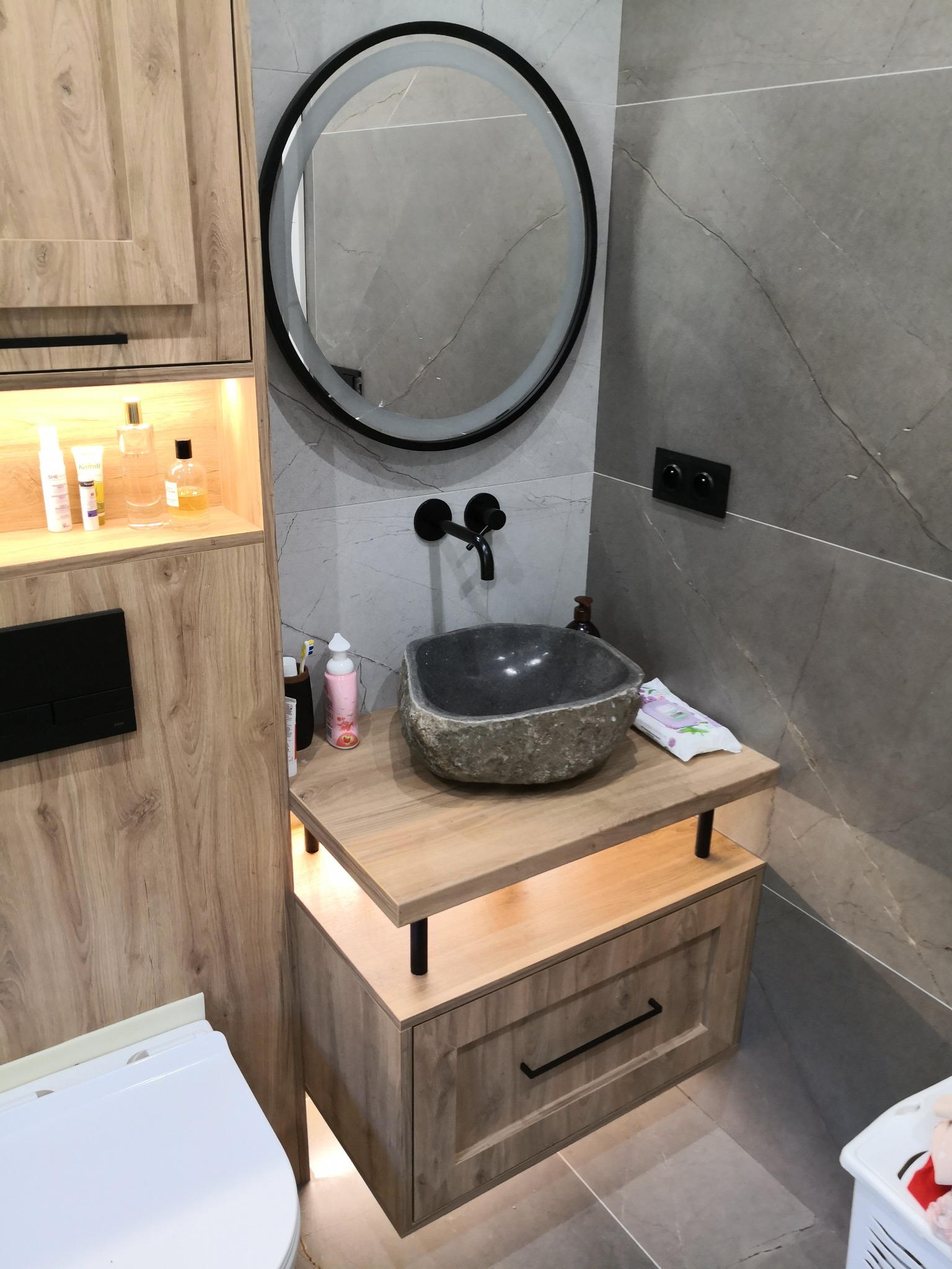 Nowoczesna łazienka z kamienną umywalką i podświetlaną szafką z drewnopodobnym wykończeniem, okrągłe lustro z podświetleniem LED i szare płytki imitujące marmur na ścianie.