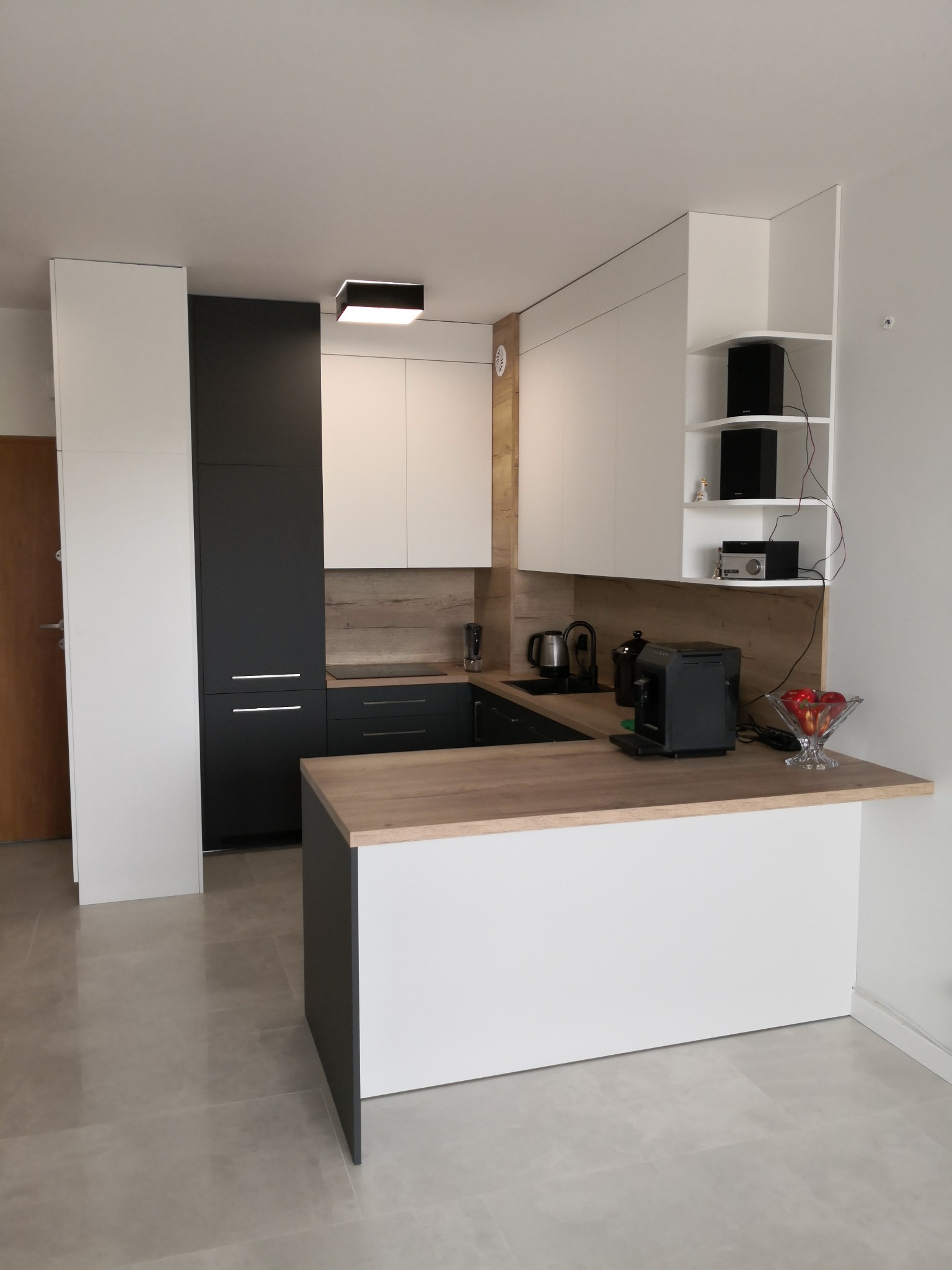 Nowoczesna kuchnia z białymi i grafitowymi frontami, blatem w kolorze jasnego drewna i wyspą, oświetlona czarną lampą sufitową.