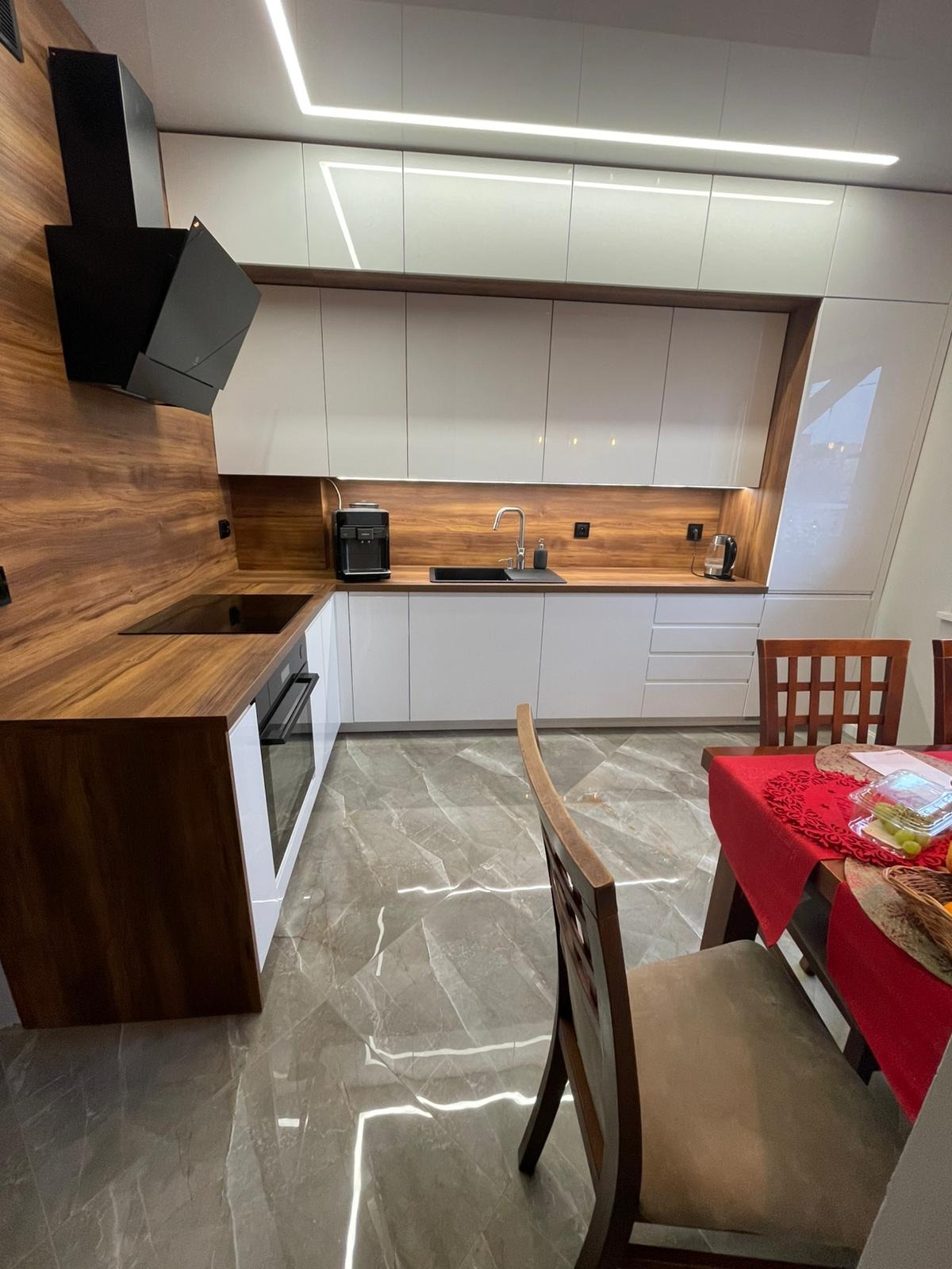 Nowoczesna kuchnia z białymi szafkami i drewnianym blatem, czarny okap nad płytą indukcyjną, oświetlenie LED podszafkowe i sufitowe, marmurowa podłoga, stół z czerwonym obrusem w tle.