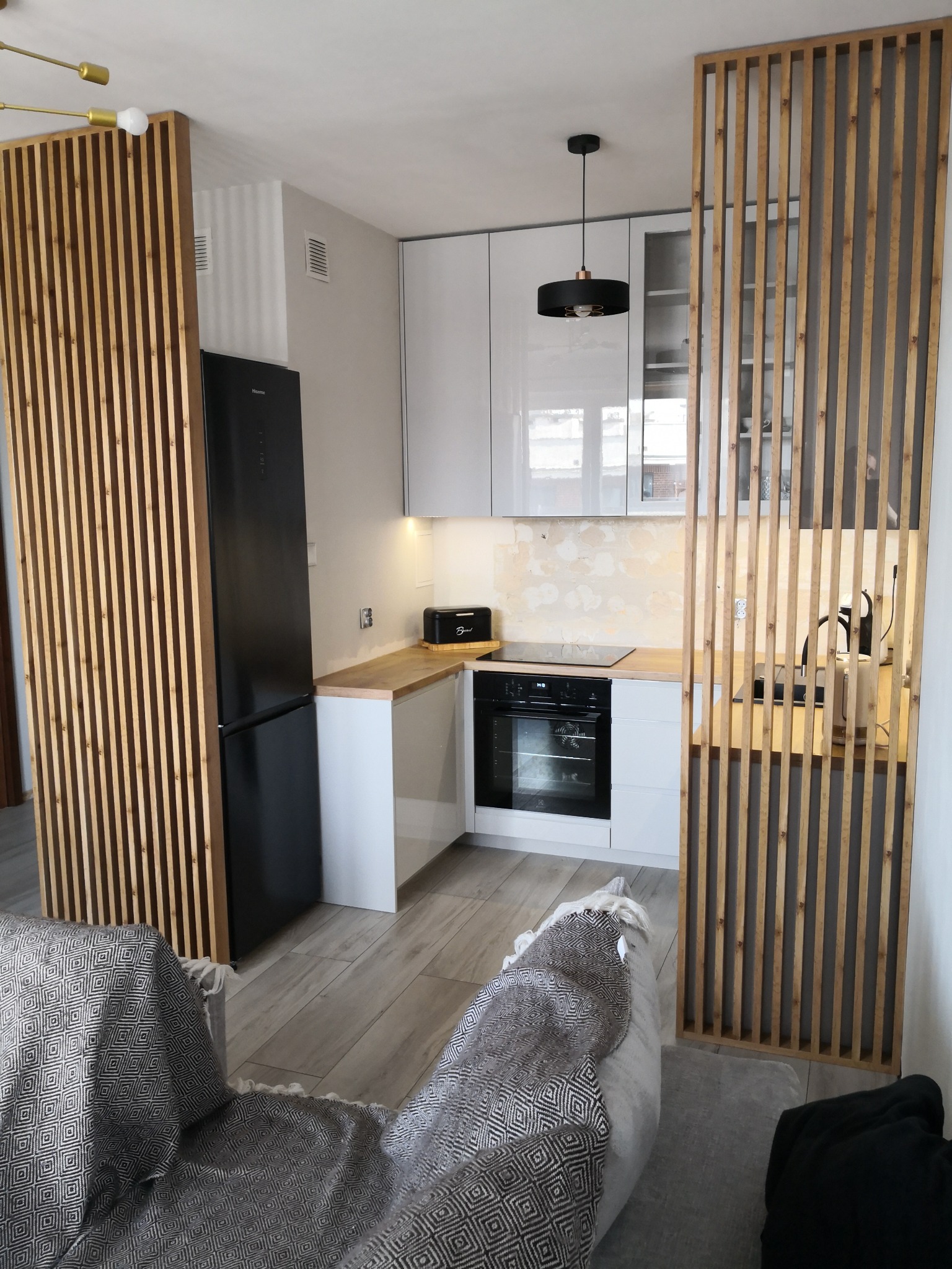 Nowoczesna kuchnia z białymi szafkami, drewnianym blatem i ażurową ścianką działową z drewnianych listew, widok z perspektywy sofy z wzorzystą narzutą.