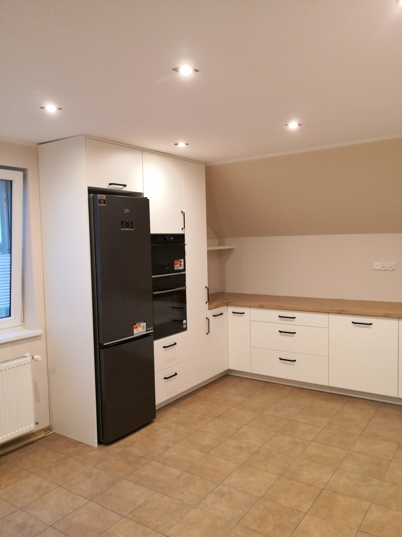 Biała zabudowa kuchenna z czarnymi uchwytami i sprzętem AGD w nowoczesnym wnętrzu. Uwagę zwraca drewniany blat i skośny sufit.