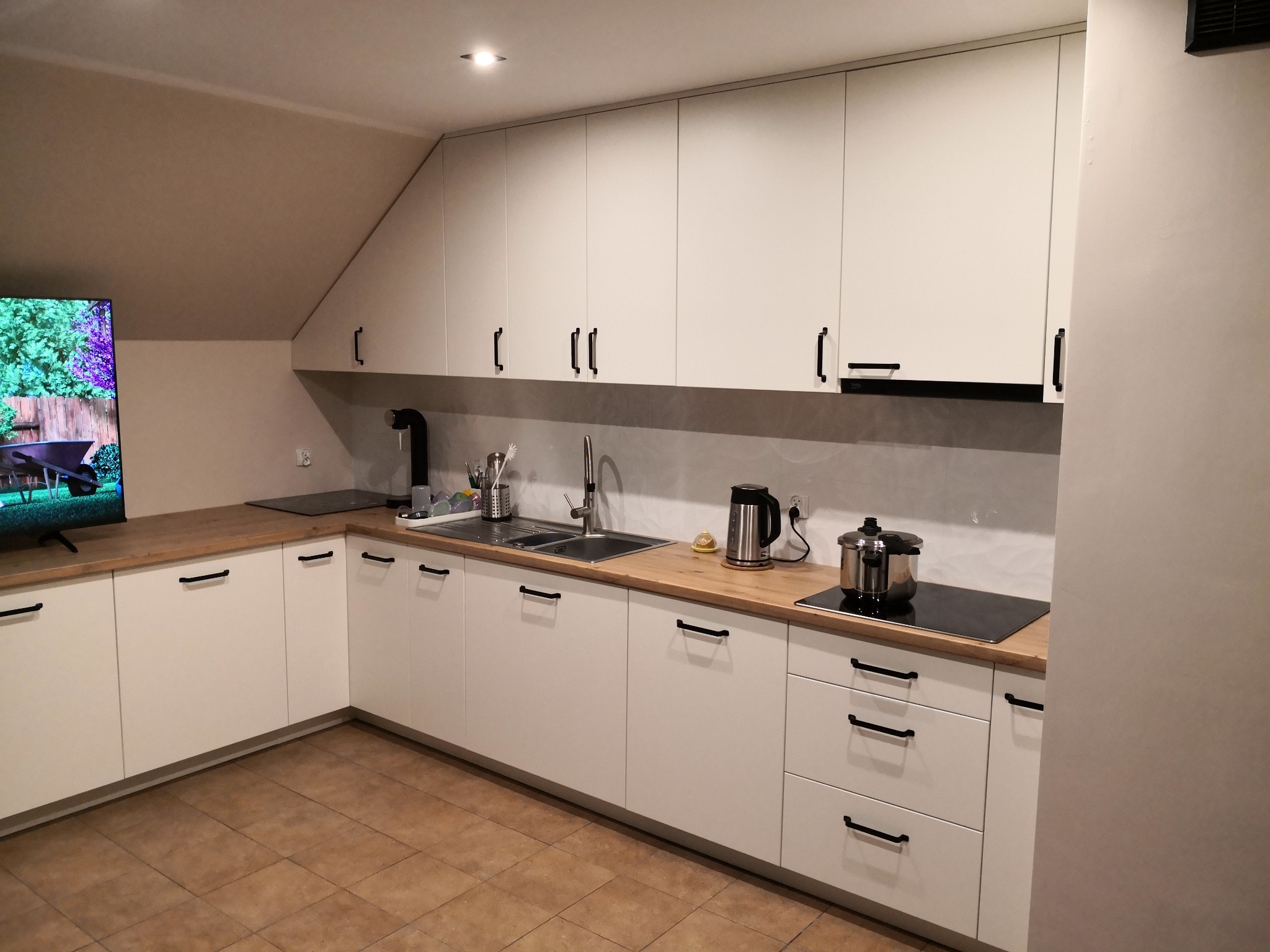 Jasna kuchnia z drewnianym blatem i czarnymi uchwytami, zlewem i płytą indukcyjną. Uwagę przykuwa telewizor z widokiem na ogród.