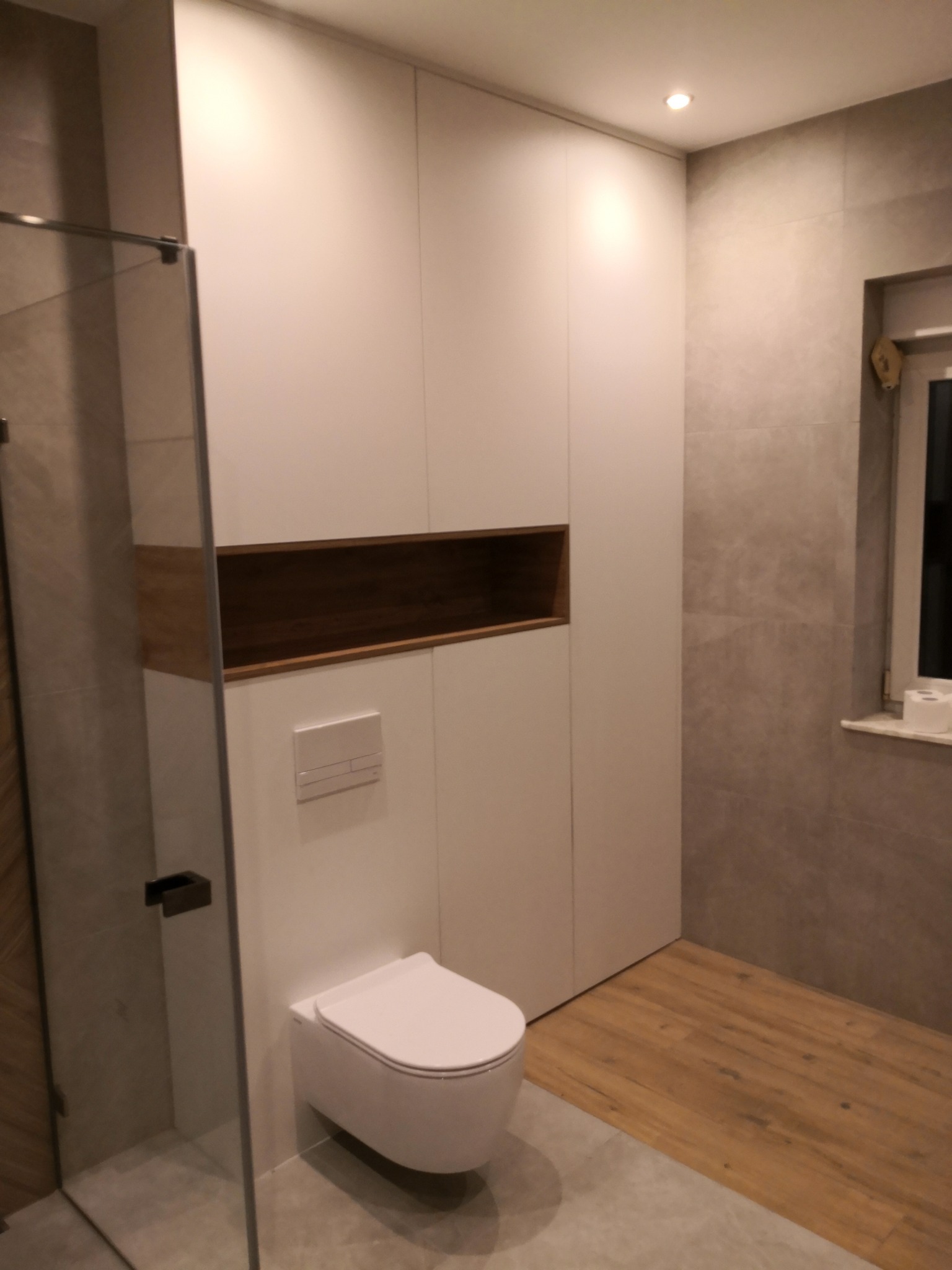 Nowoczesna łazienka z białą szafą wnękową z drewnianą wnęką, podwieszaną toaletą i kabiną prysznicową. Szare płytki i drewnopodobna podłoga.