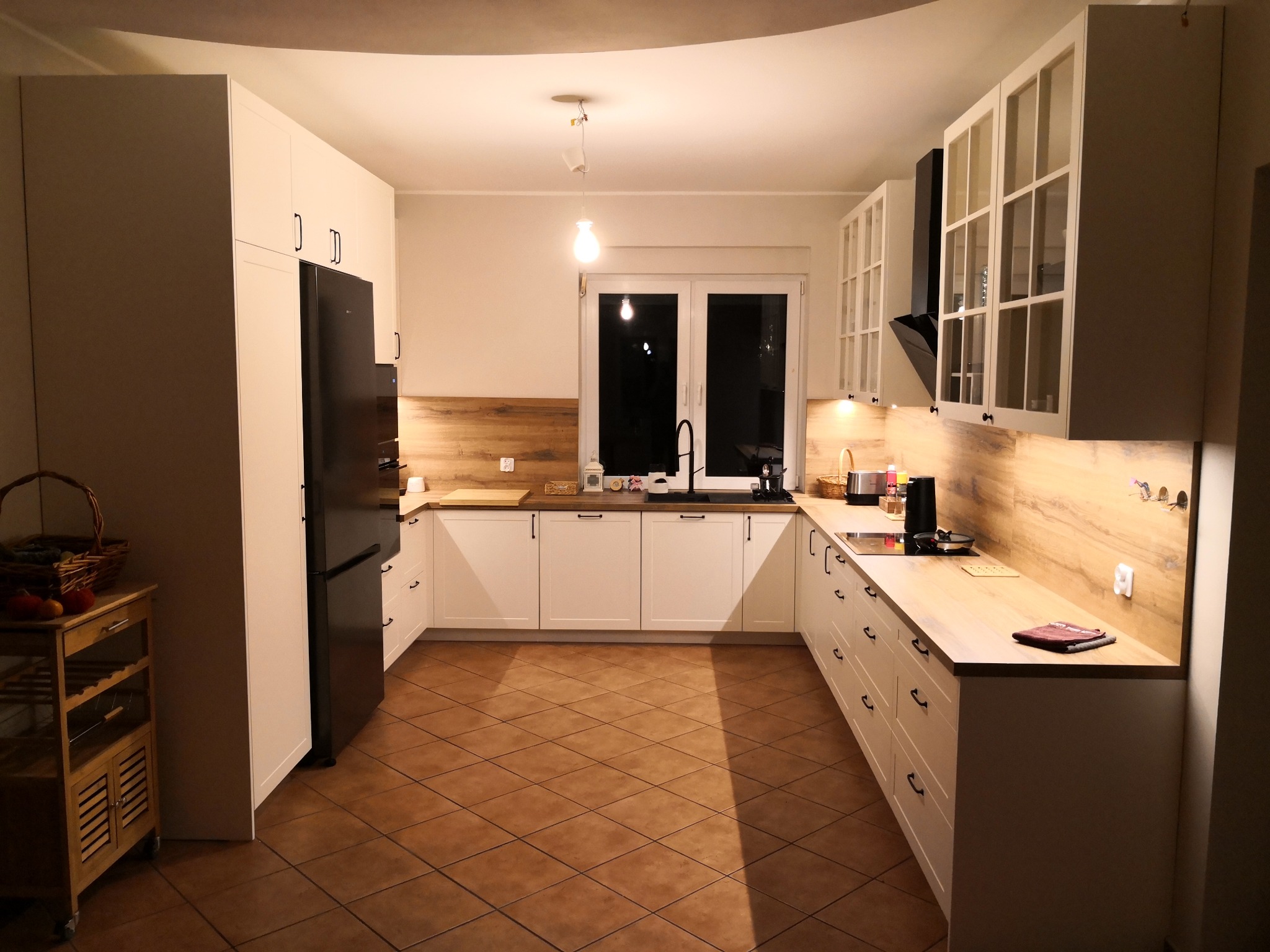 Jasna kuchnia z drewnianym blatem i czarnymi uchwytami. Uwagę przykuwają przeszklone szafki wiszące i czarna lodówka side-by-side. Ciepłe światło podkreśla przytulny charakter wnętrza.