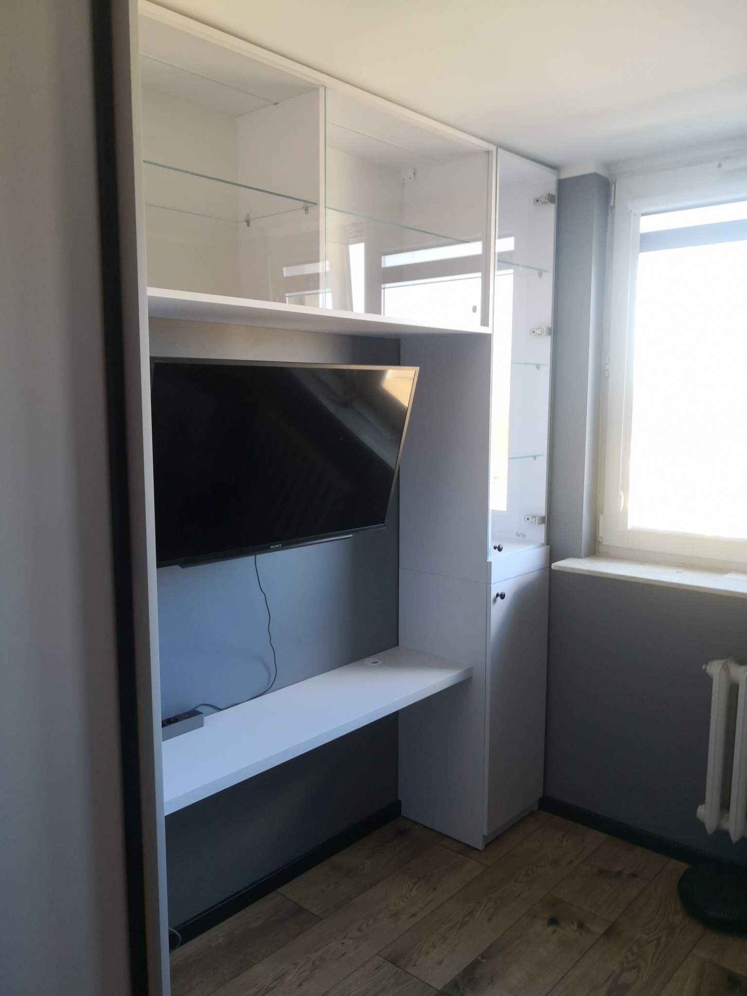 Biała szafa wnękowa z szklanymi półkami i miejscem na telewizor, uzupełniona blatem biurka. Nowoczesny design, drewniana podłoga, szara ściana.