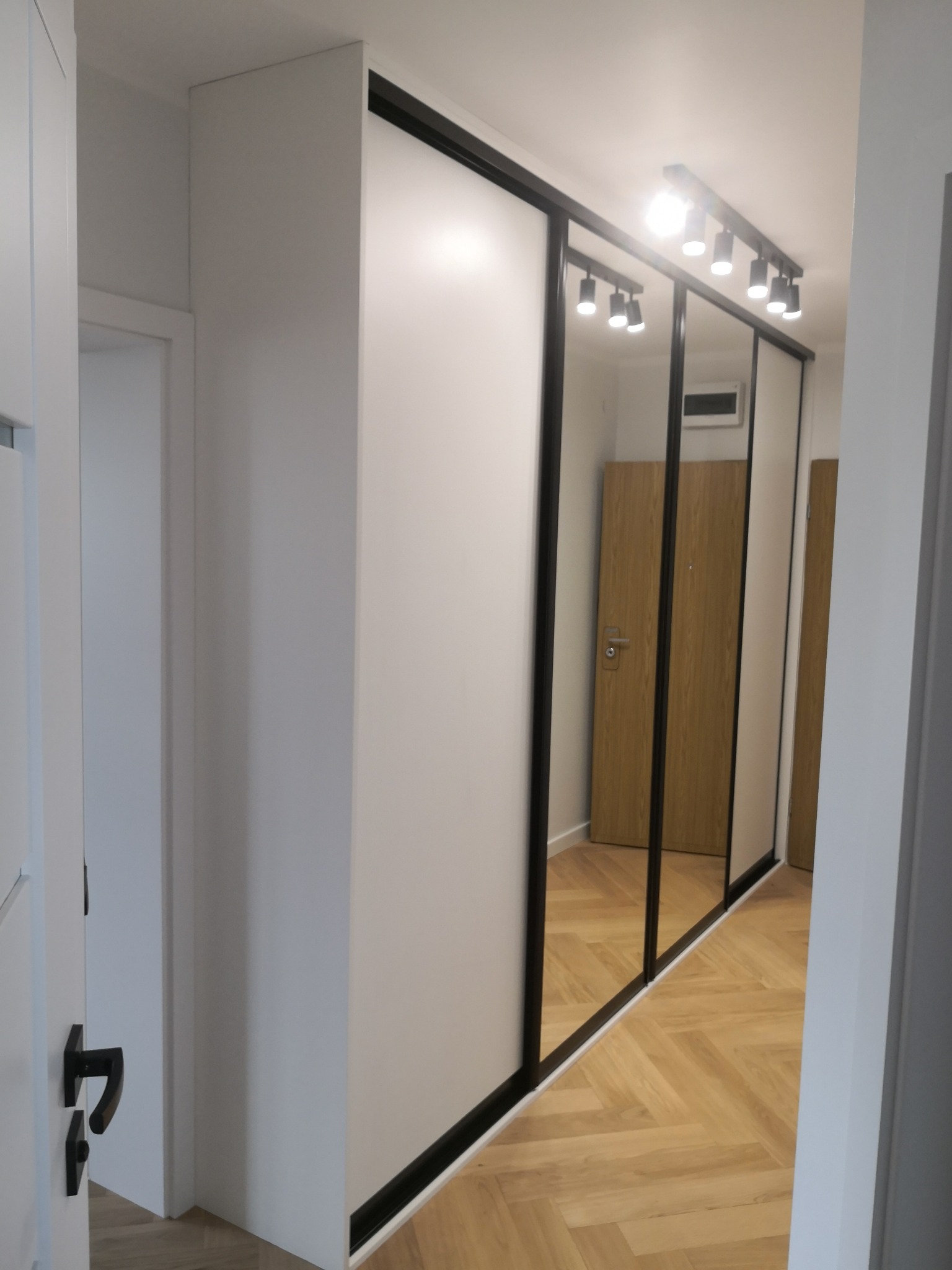 Biała szafa wnękowa z przesuwnymi drzwiami z czarnymi ramami i lustrami, oświetlona punktowo, na podłodze parkiet w jodełkę. Widok z perspektywy korytarza.