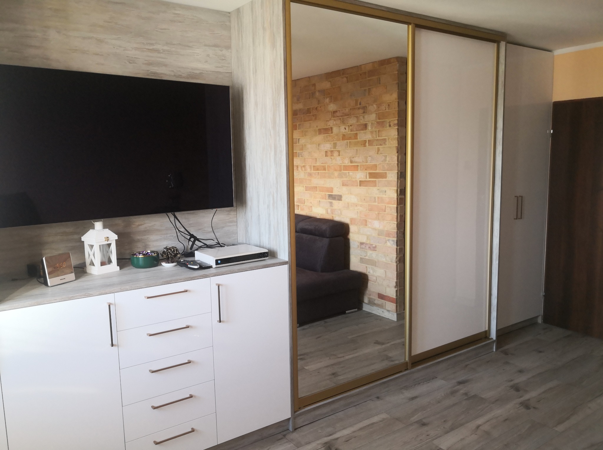 Biała szafa wnękowa z lustrzanymi drzwiami przesuwnymi, zintegrowana z szafką RTV. Widoczny fragment ceglanej ściany w odbiciu lustra.