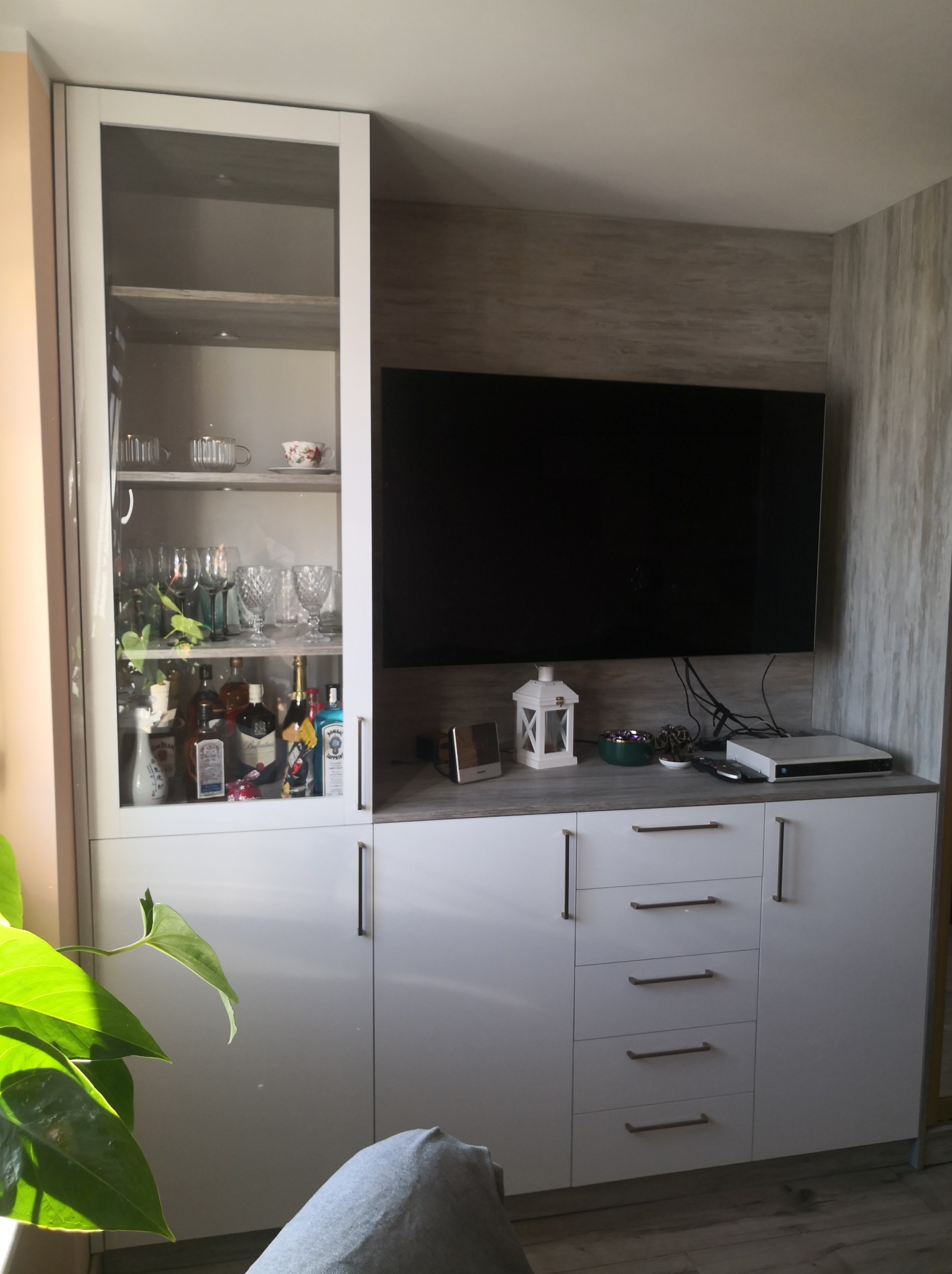 Biała zabudowa meblowa z witryną i szafkami pod telewizor. Na półkach szklanki i butelki. Minimalistyczne uchwyty. Część rośliny w kadrze.