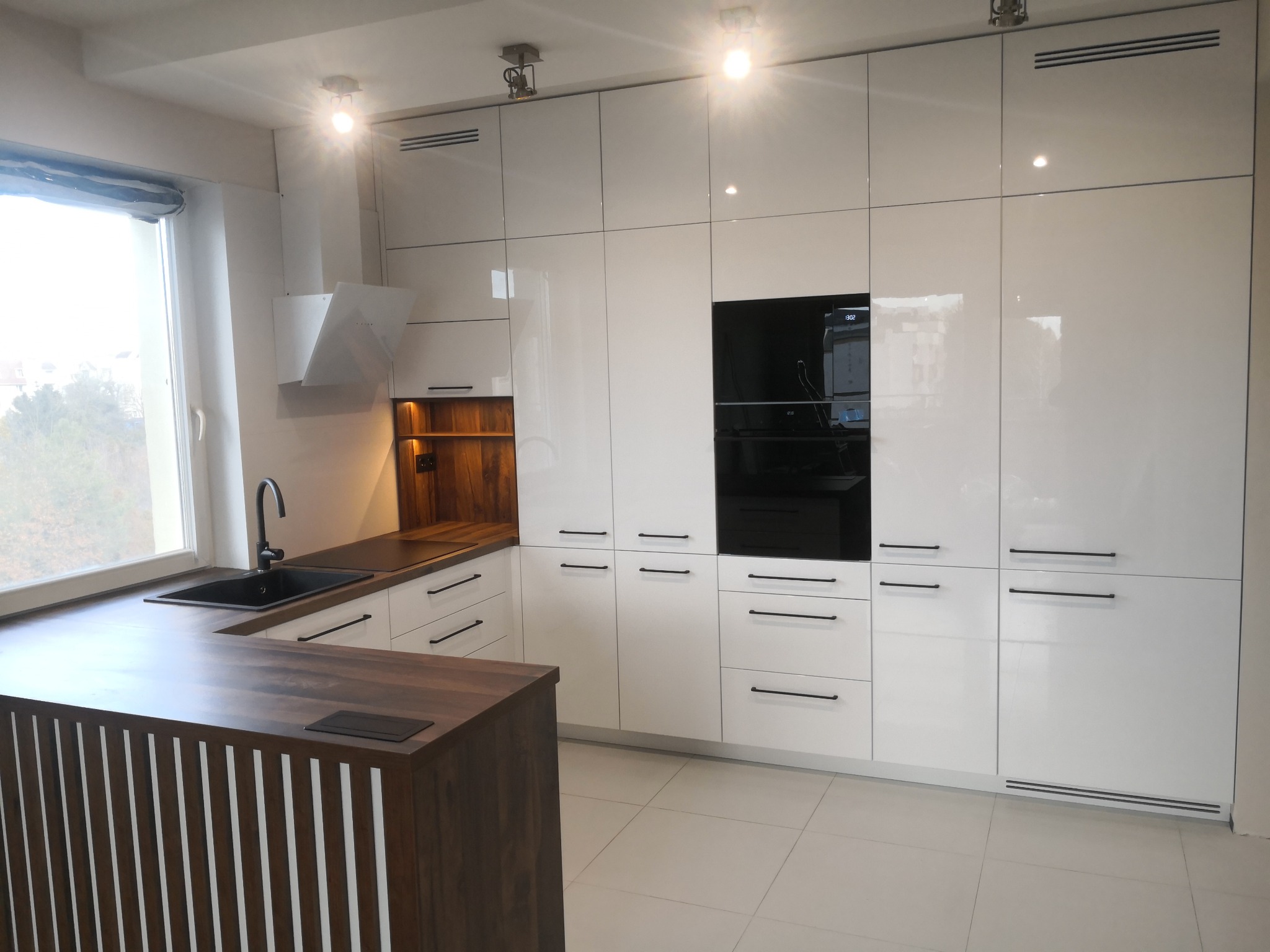 Biała, nowoczesna kuchnia z drewnianym blatem i wyspą. Fronty szafek na wysoki połysk, czarne, minimalistyczne uchwyty. Widoczny okap i zabudowany sprzęt AGD.