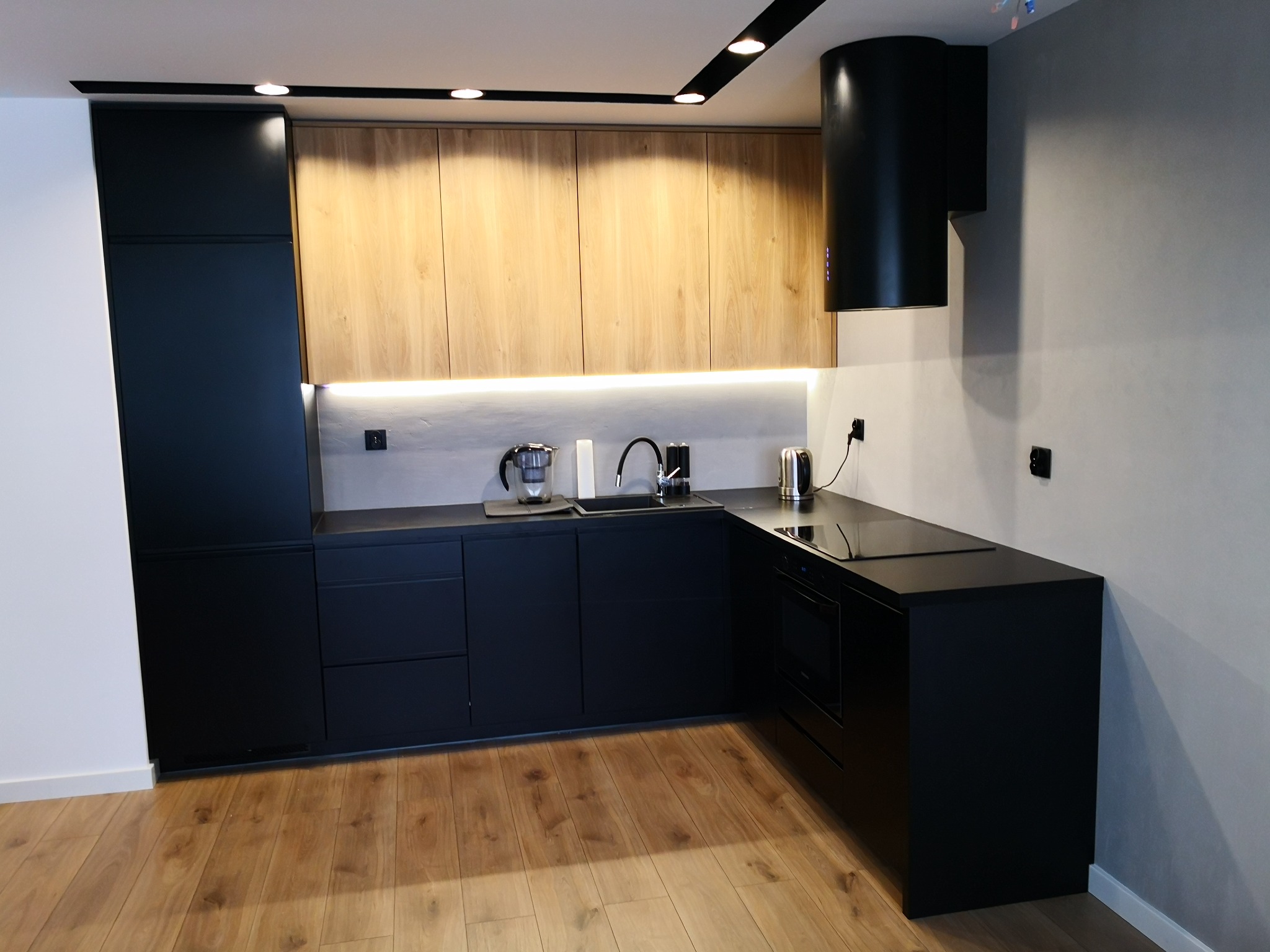 Nowoczesna kuchnia z czarnymi szafkami dolnymi i drewnianymi górnymi, oświetlona listwą LED pod szafkami, z czarnym okapem cylindrycznym.