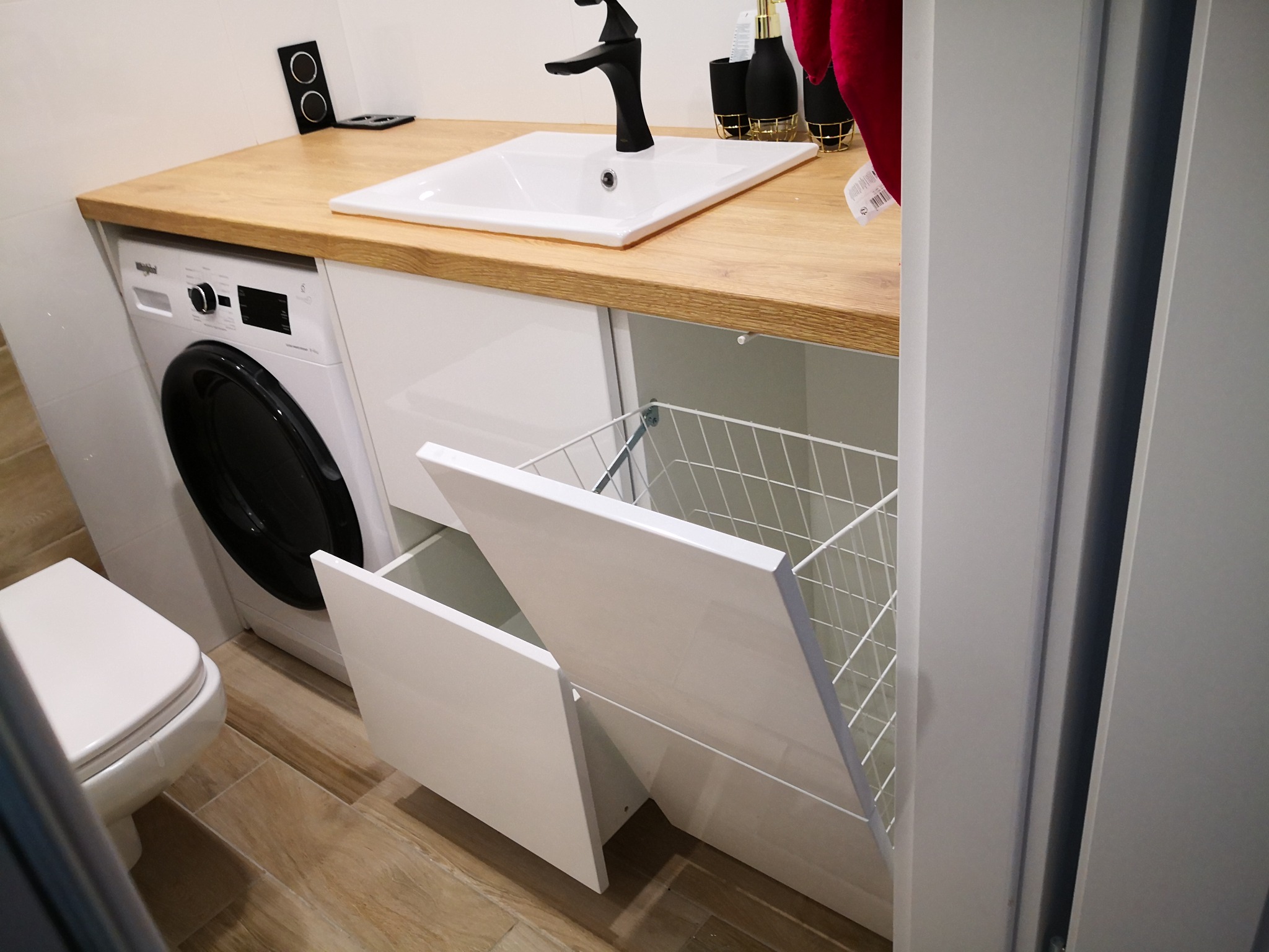 Nowoczesna łazienka z drewnianym blatem, białą umywalką i czarną armaturą, pralką w zabudowie oraz wysuwanym koszem na bieliznę.