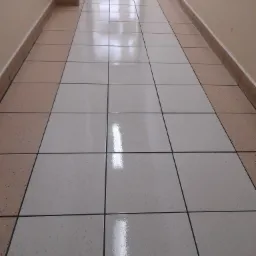 Długi korytarz wyłożony jasnymi i beżowymi płytkami podłogowymi z ciemnymi fugami, widziany z perspektywy podłogi, z odbiciem światła na powierzchni.