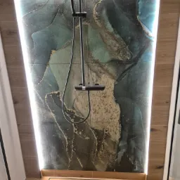 Kabina prysznicowa z deszczownicą i panelami ściennymi z efektem marmuru, podświetlona listwą LED, z odpływem liniowym w podłodze wyłożonej płytkami imitującymi drewno.