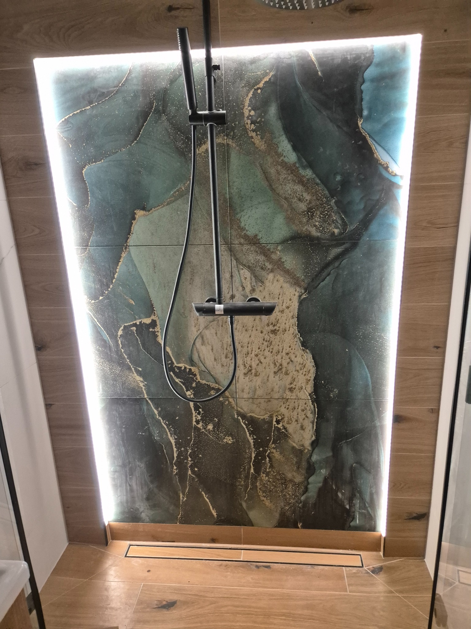 Kabina prysznicowa z deszczownicą i panelami ściennymi z efektem marmuru, podświetlona listwą LED, z odpływem liniowym w podłodze wyłożonej płytkami imitującymi drewno.