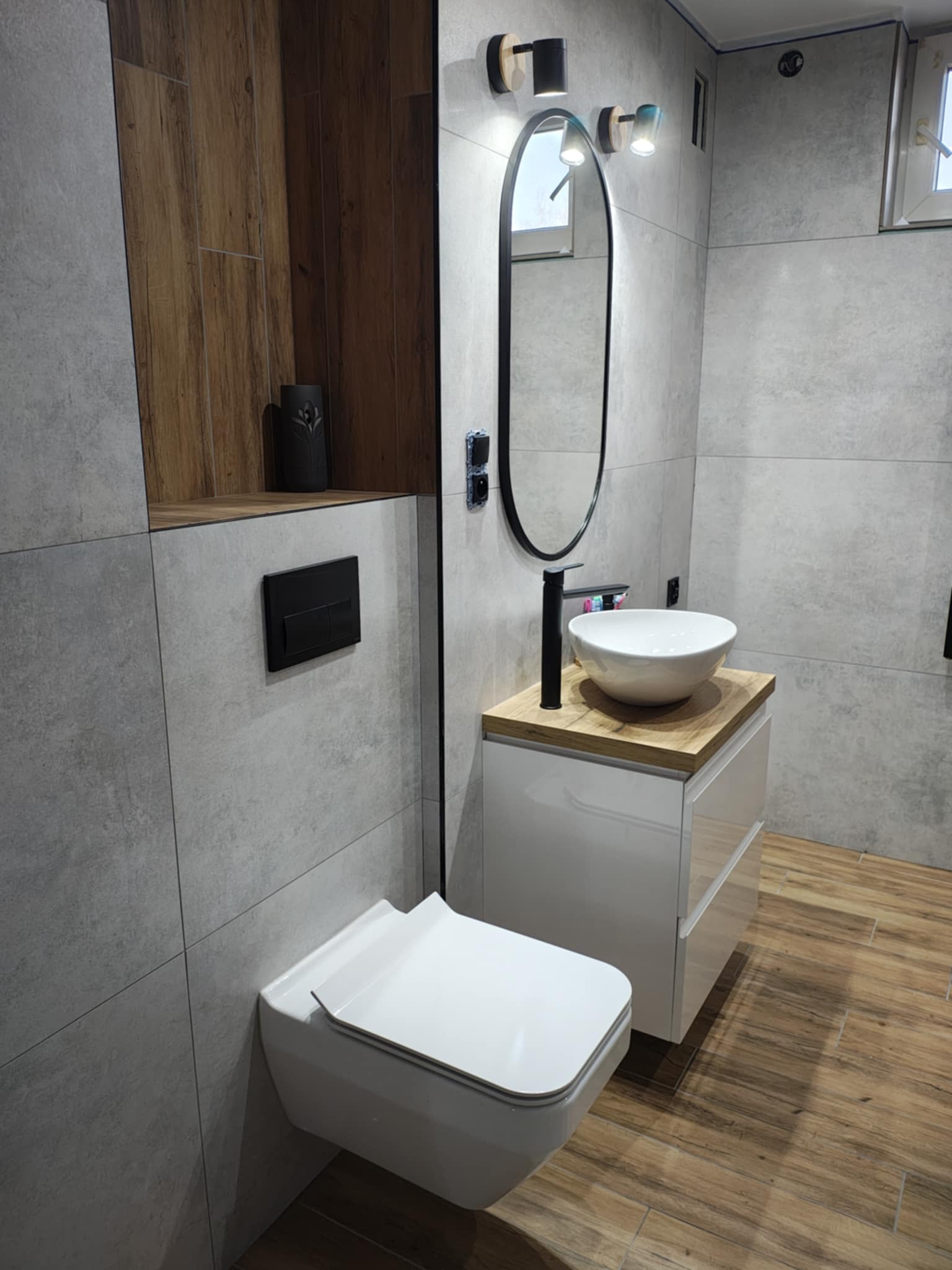 Nowoczesna łazienka w Chabsku z szarą płytką imitującą beton, drewnianymi akcentami, owalnym lustrem z podświetleniem LED, wiszącą miską WC i umywalką nablatową na szafce.