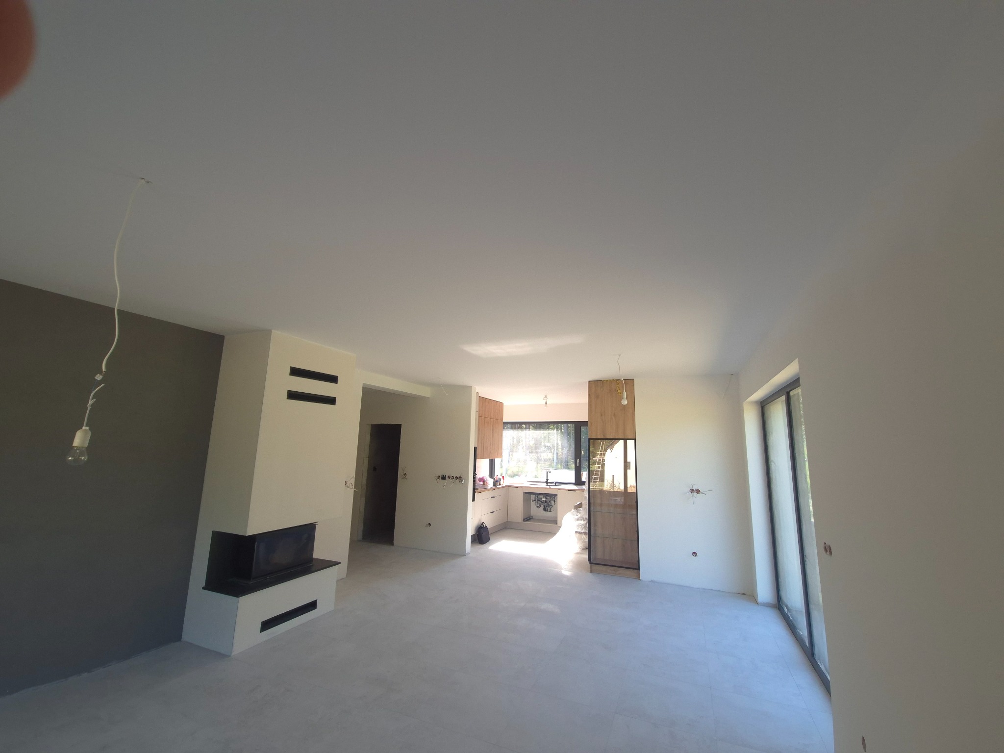 Jasne, przestronne wnętrze po remoncie: szare ściany, kominek z czarnymi akcentami, okno przesuwne i widok na kuchnię. Minimalistyczny design i neutralna kolorystyka.