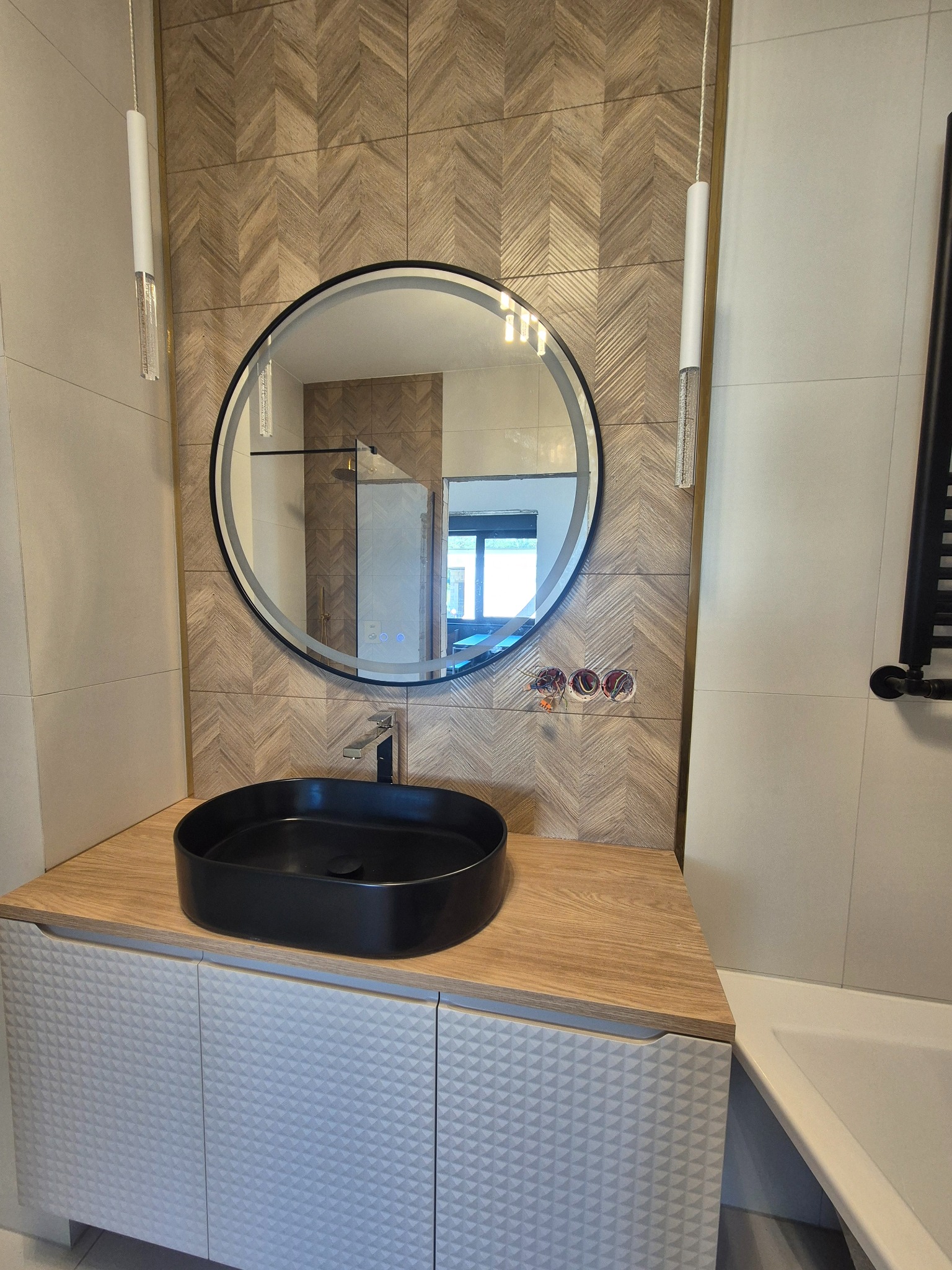 Nowoczesna aranżacja łazienki: czarna umywalka na szafce z geometrycznym wzorem, okrągłe lustro z podświetleniem i dekoracyjne płytki imitujące drewno na ścianie.