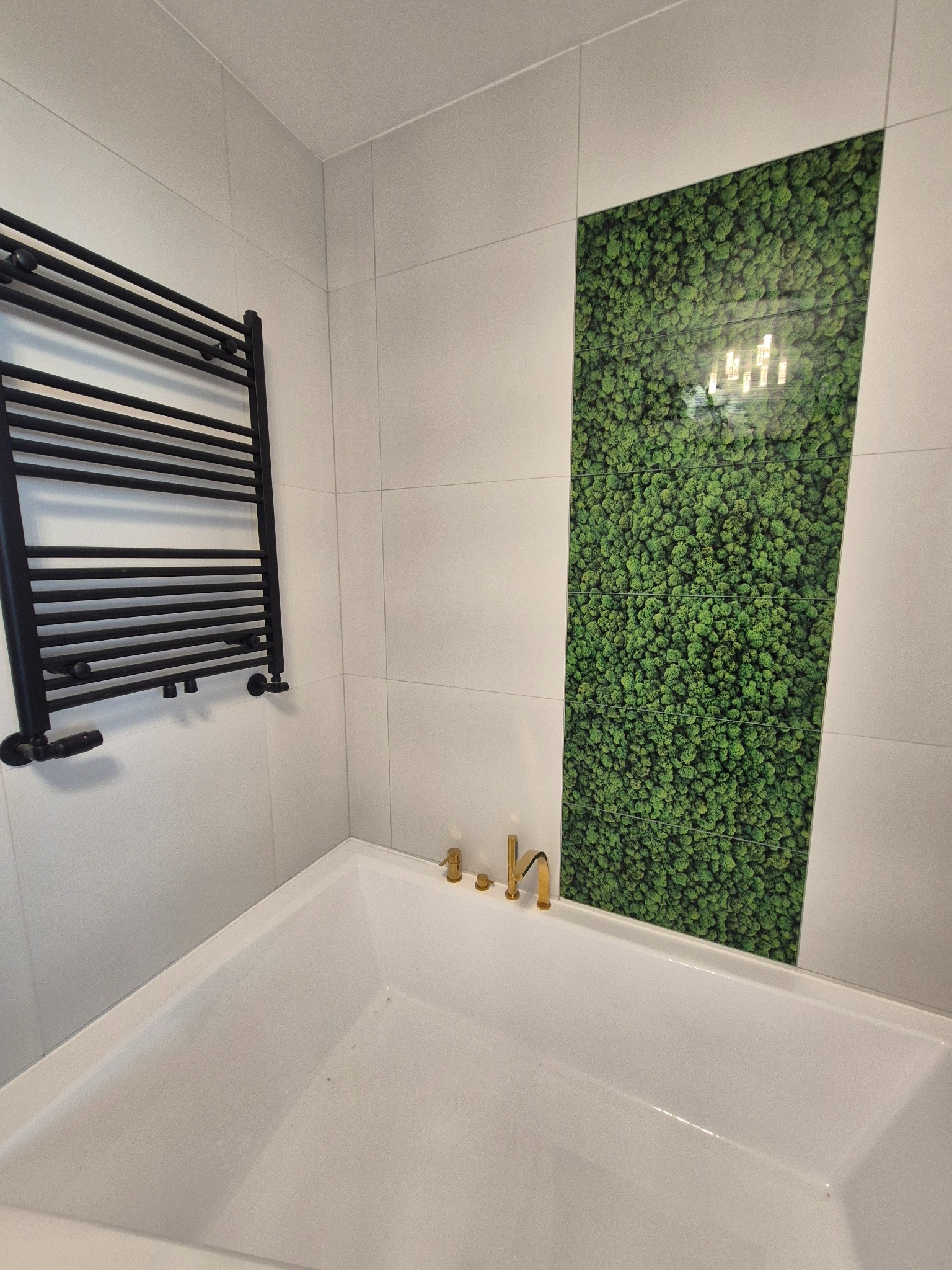 Nowoczesna łazienka z białą wanną, czarnym grzejnikiem i zielonym panelem z mchu na ścianie. Złota bateria umywalkowa dodaje elegancji.