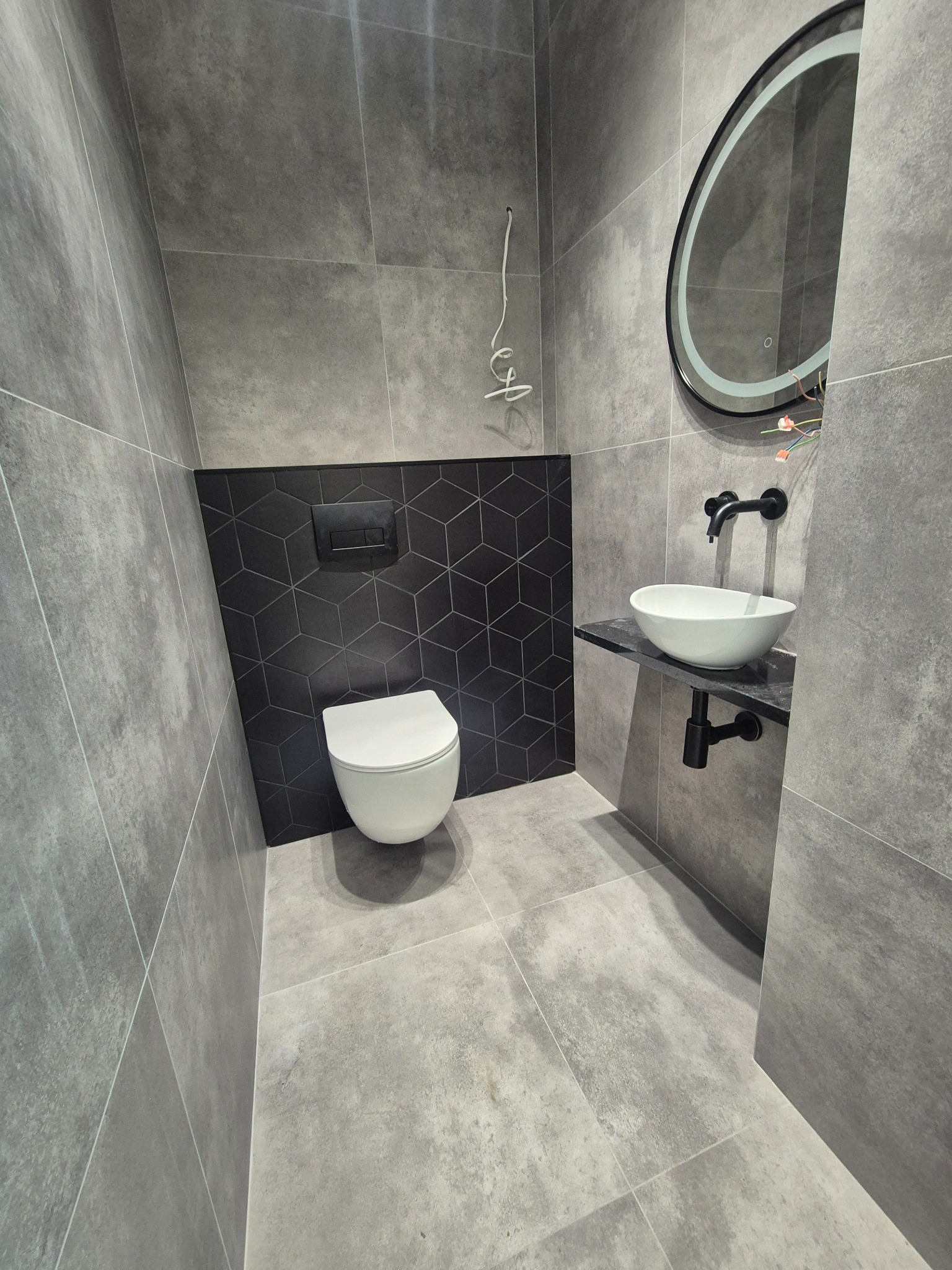 Nowoczesna łazienka w odcieniach szarości z czarnymi akcentami: umywalka nablatowa, podświetlane lustro i toaleta. Na ścianie geometryczne, czarne płytki.
