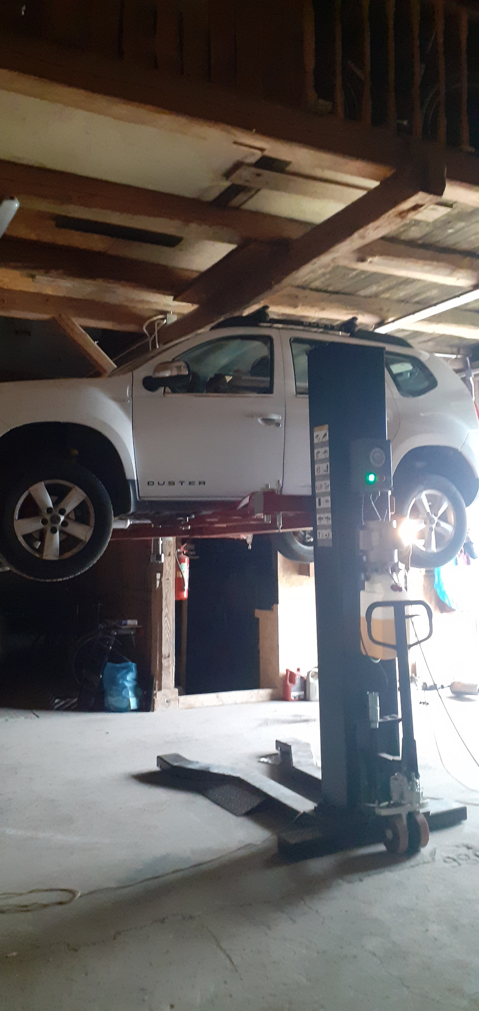 Biały samochód marki Duster na podnośniku w warsztacie, widoczne otwarte drzwi i podniesiona maska, w tle drewniana konstrukcja dachu.