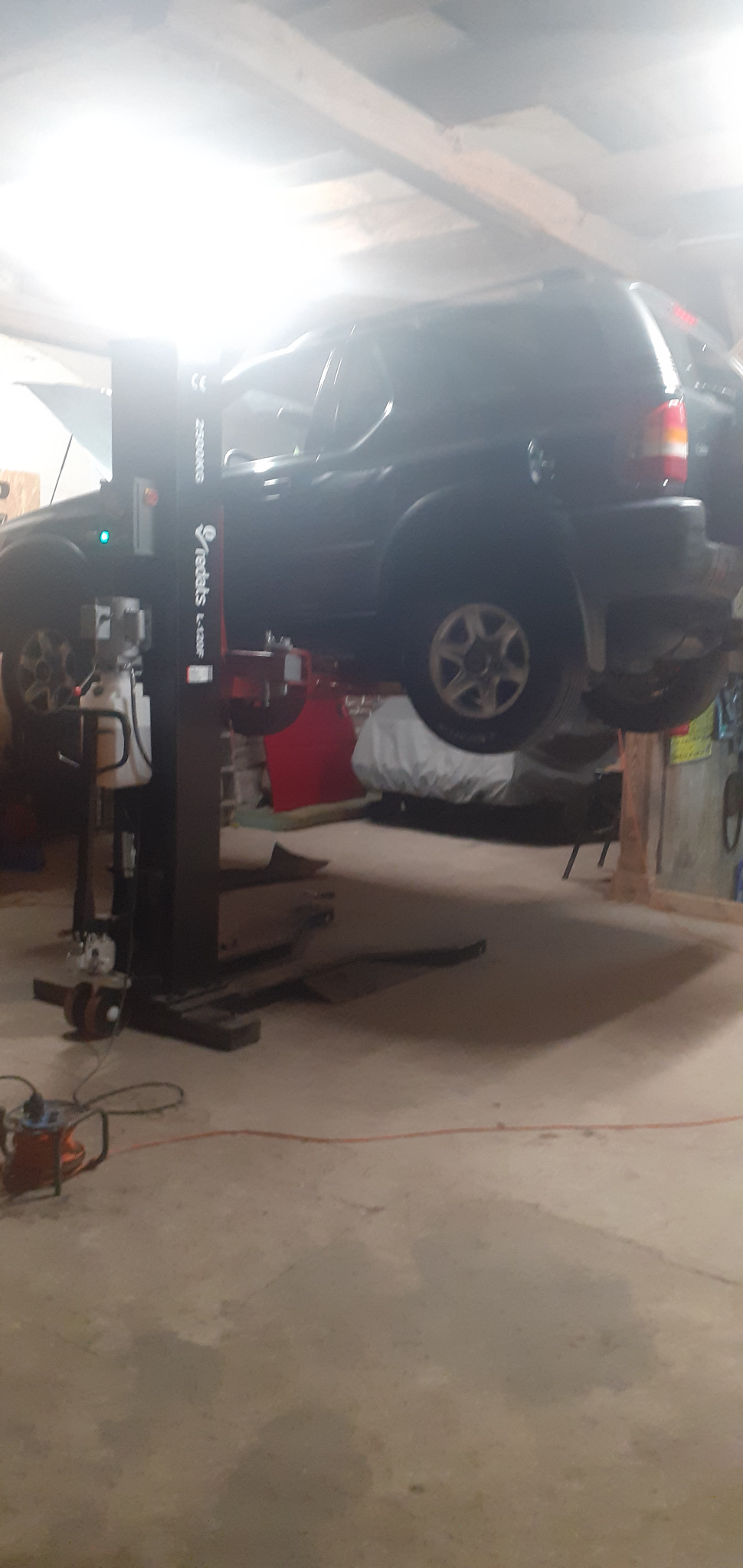 Czarny SUV podniesiony na dwukolumnowym podnośniku Redats w warsztacie, widoczny przewód od spawarki i podłoga z plamami.