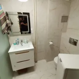 Wykończona łazienka z białymi marmurkowymi płytkami, białą szafką pod umywalką i pralką, widoczne odbicie fotografa w lustrze.