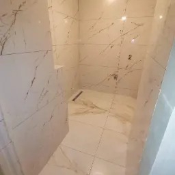 Płytki imitujące marmur z subtelnymi złotymi żyłami w kabinie prysznicowej, widoczny odpływ liniowy i przygotowane punkty wodne na ścianie.