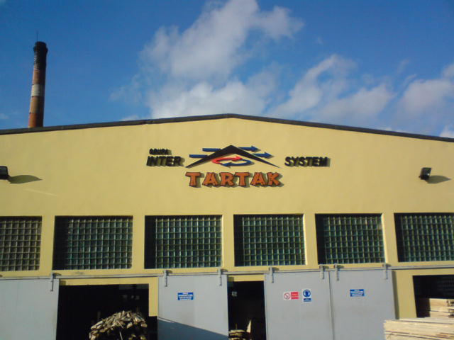 Budynek tartaku 'Inter System' z widocznym kominem w tle, fragment składowanego drewna przed budynkiem.