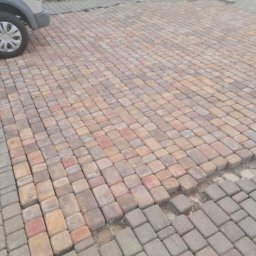 Zabdziobud - Fragment nawierzchni z kostki brukowej w odcieniach czerwieni i szarości, z widocznymi brakami w ułożeniu i stertą kostki w tle, częściowo zasłoniętą przez samochód.