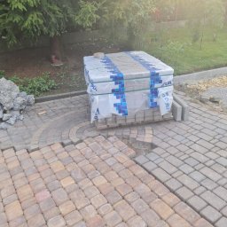 Zabdziobud - Fragment układanej nawierzchni z kostki w odcieniach brązu i szarości, obok paleta z zapakowaną kostką oraz pryzma piasku, w tle zieleń ogrodu.