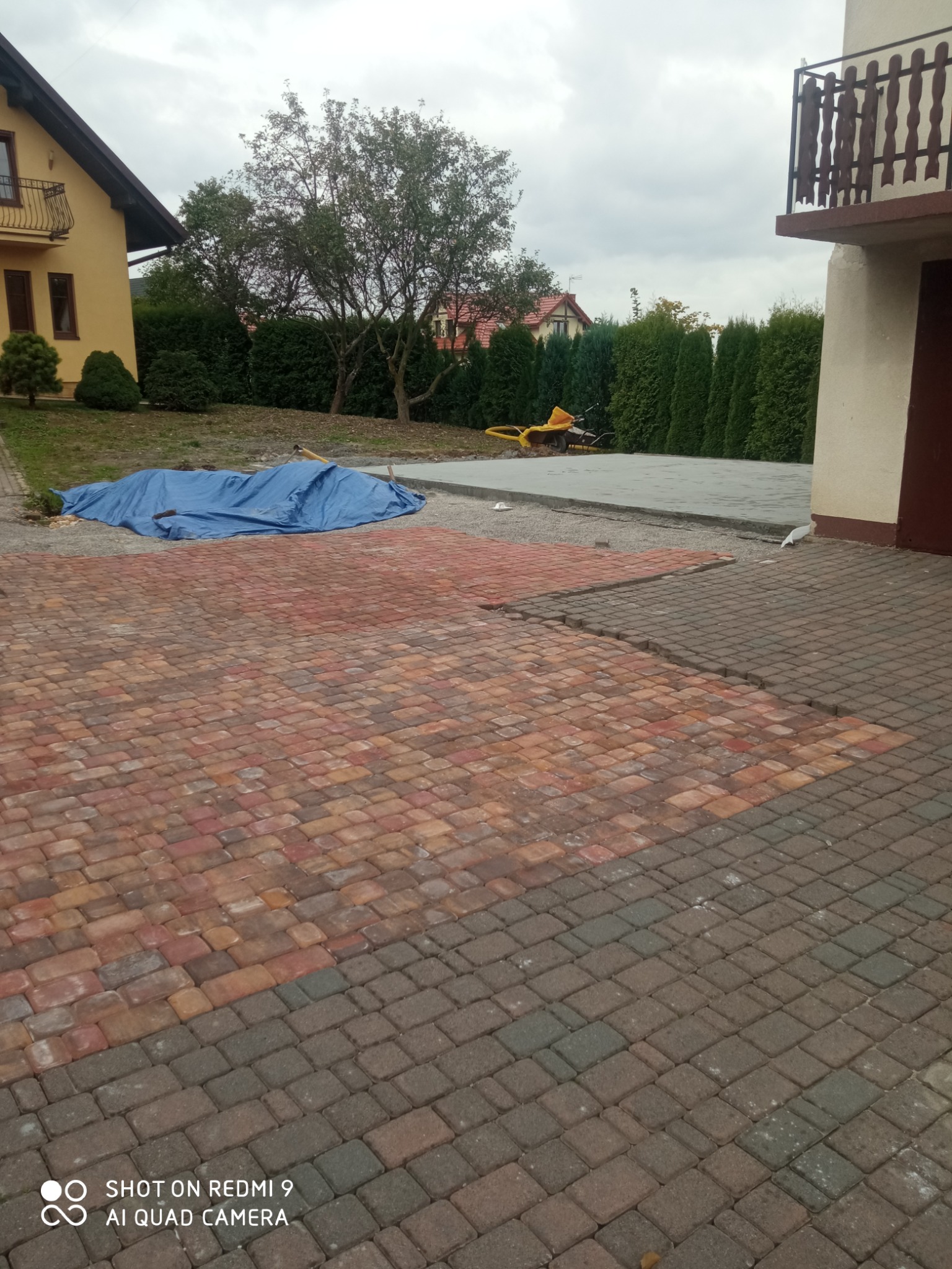 Fragment posesji w trakcie prac brukarskich: ułożona część z czerwono-brązowej kostki, przygotowany podkład pod kolejną partię, narzędzia i materiały budowlane pod plandeką w tle.