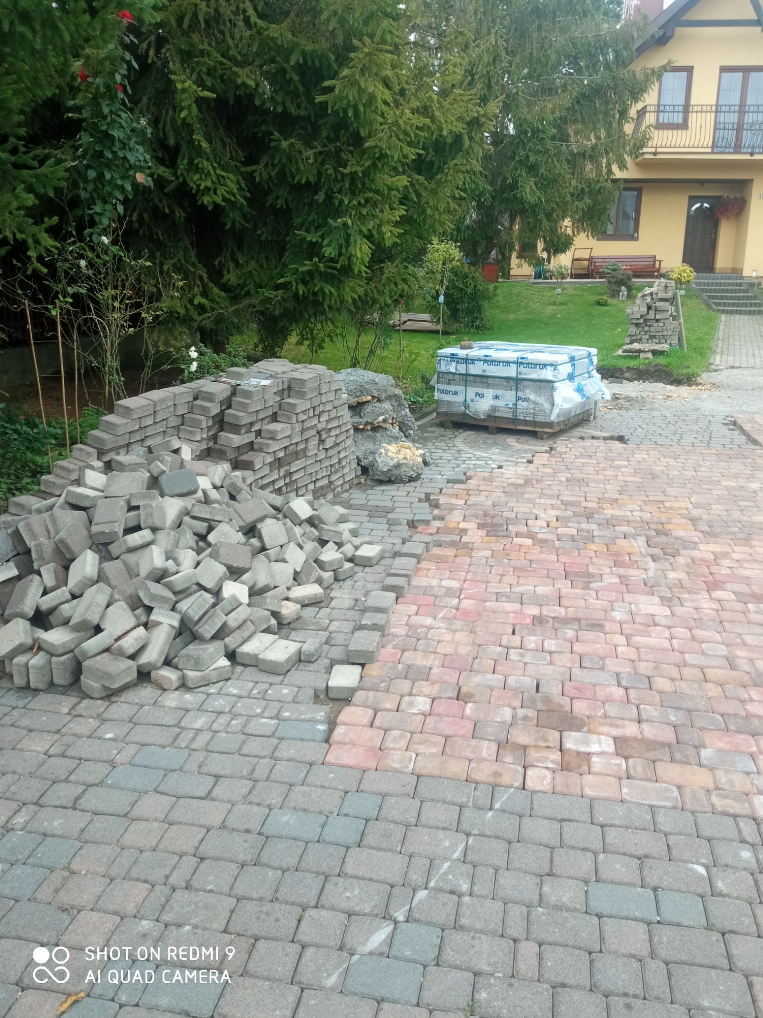 Stos szarych kostek brukowych obok ułożonej nawierzchni z kostki w odcieniach czerwieni i szarości, w tle dom z zielonym ogrodem i paleta zafoliowanych kostek.