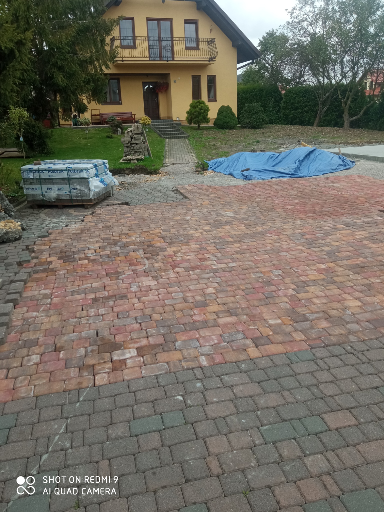 Częściowo ułożona nawierzchnia z kostki w odcieniach czerwieni i szarości przed domem z żółtą elewacją, na palecie zapakowane kostki brukowe.