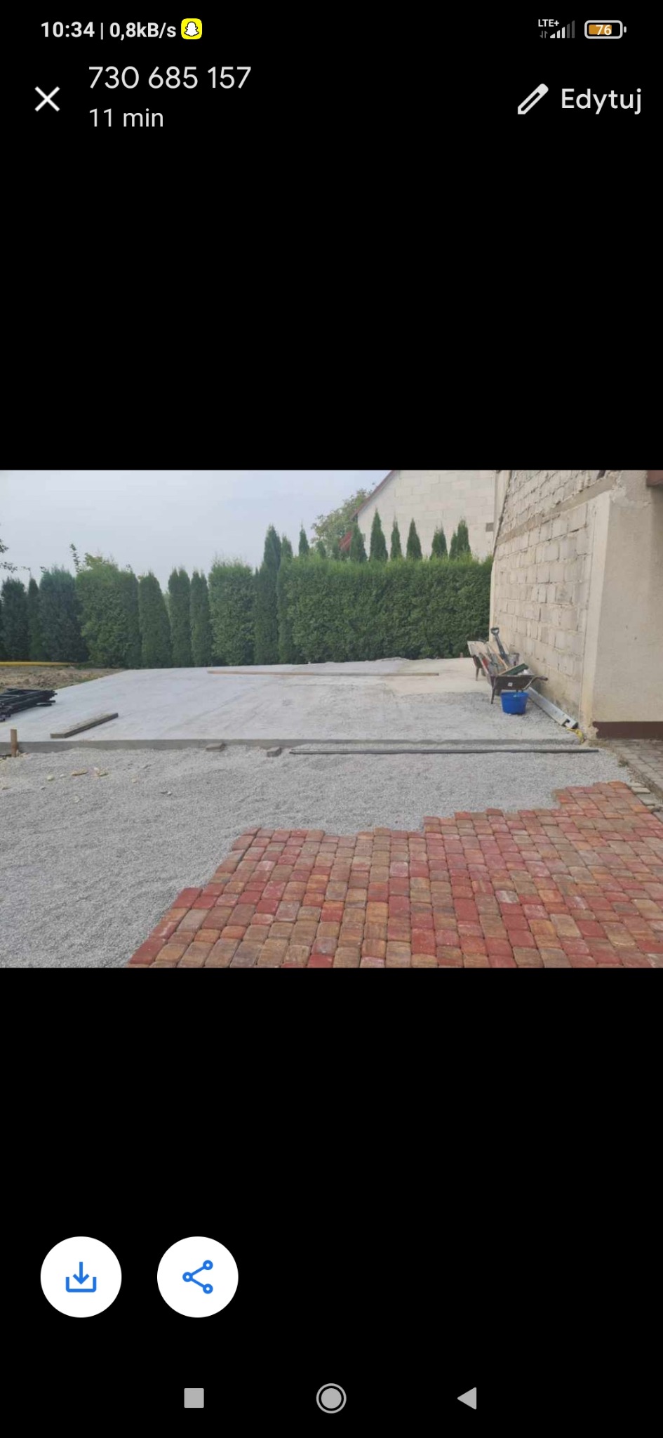 Fragment układanej nawierzchni z czerwono-brązowej kostki, widoczny przygotowany podkład z kruszywa, w tle rząd tuj i ściana budynku z narzędziami.