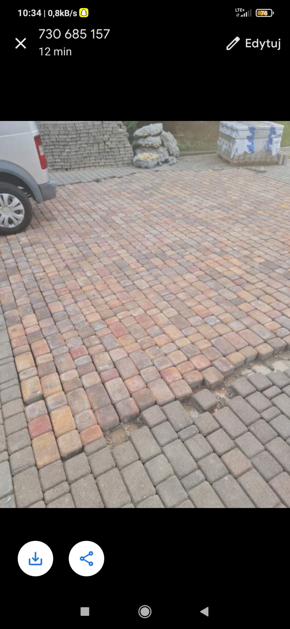 Fragment nawierzchni z kostki brukowej w odcieniach czerwieni i szarości, z widocznymi brakami w ułożeniu i stertą kostki w tle, częściowo zasłoniętą przez samochód.