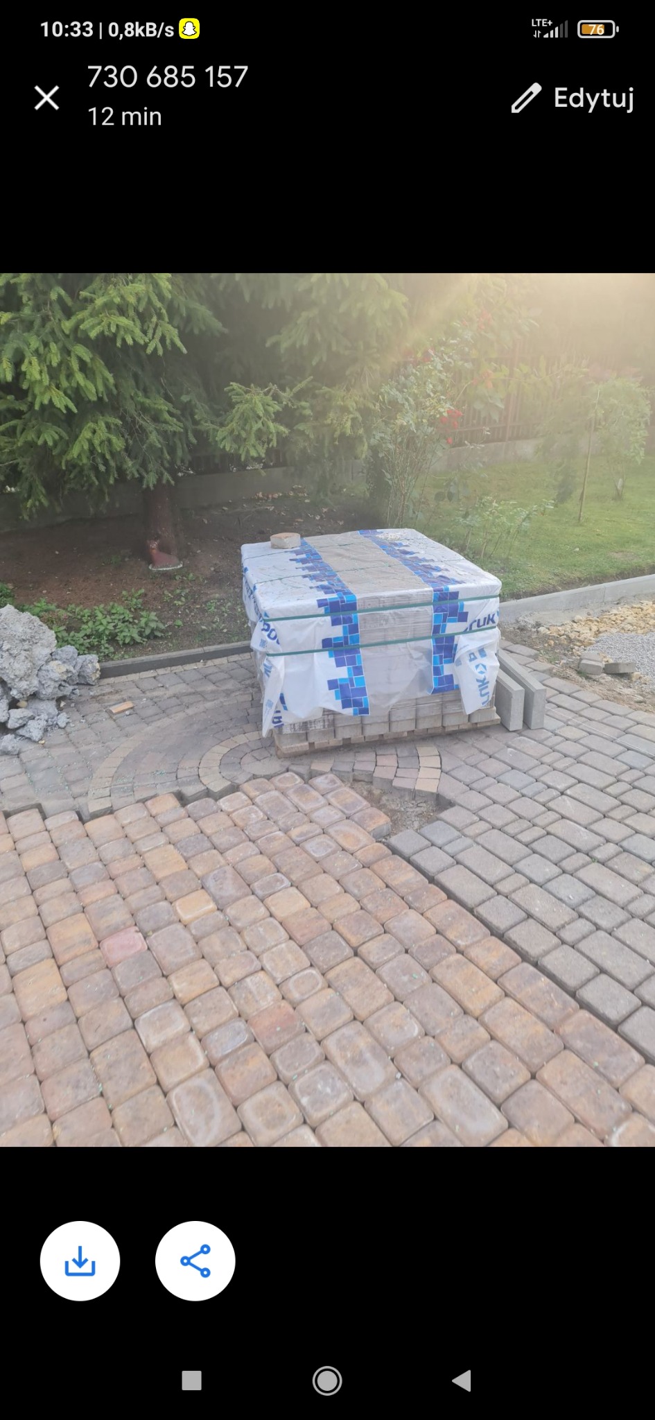 Fragment układanej nawierzchni z kostki w odcieniach brązu i szarości, obok paleta z zapakowaną kostką oraz pryzma piasku, w tle zieleń ogrodu.