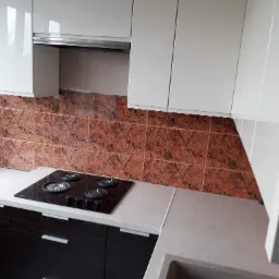 Fragment kuchni z białymi szafkami wiszącymi, brązowymi płytkami na ścianie i czarną płytą grzewczą, widoczne niedokończone wykończenie ściany przy oknie.