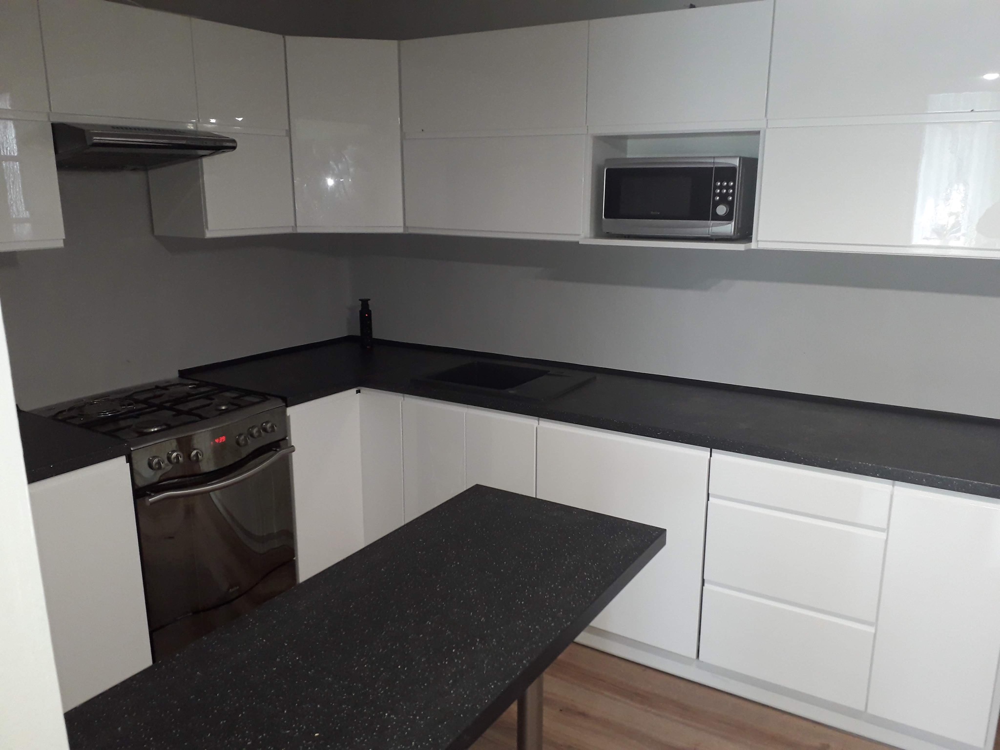 Nowoczesna kuchnia z białymi, lakierowanymi frontami, czarnym blatem z drobinkami i wysuwana wyspa na metalowej nodze, widoczna kuchenka gazowa i mikrofalówka.