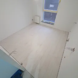 Jasne panele podłogowe ułożone w pokoju, okno zabezpieczone taśmą malarską, widoczne wystające kable elektryczne ze ściany.