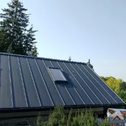 Nowoczesny, ciemny dach z blachy na rąbek stojący z oknem dachowym, na tle zieleni i błękitnego nieba. Widoczny fragment rynny i elewacji budynku.