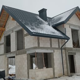 Km Dachy - Dom w stanie surowym otwartym, z częściowo wykonanym dachem z blachodachówki w kolorze grafitowym i zamontowaną rynną. Widoczny komin i śnieg.