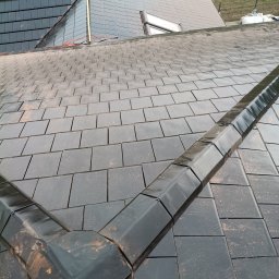 Km Dachy - Czarny dach z dachówki ceramicznej, widok z góry na fragment połaci z rynną koszową. W tle fragment dachu z oknem połaciowym i zielone pole.