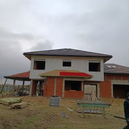 Km Dachy - Budowa domu jednorodzinnego w trakcie realizacji. Widoczny fragment dachu z brązową dachówką, ściany z czerwonej cegły i pracownik w ubraniu roboczym.