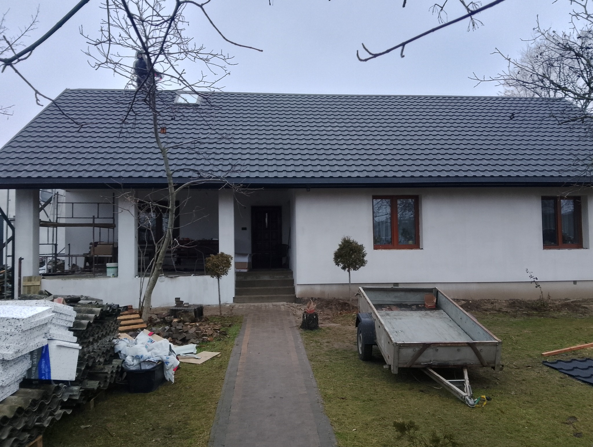 Dom z nowym, grafitowym dachem z blachodachówki. Na dachu widoczna postać. Na pierwszym planie materiały budowlane i przyczepka. Ujęcie z dołu, pochmurne niebo.