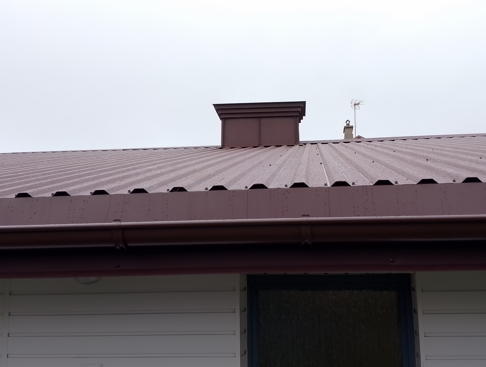 Ciemnobrązowy dach z blachy trapezowej, z widocznymi elementami wykończenia i rynnami w tym samym kolorze. W tle komin i antena telewizyjna. Fragment ściany budynku.